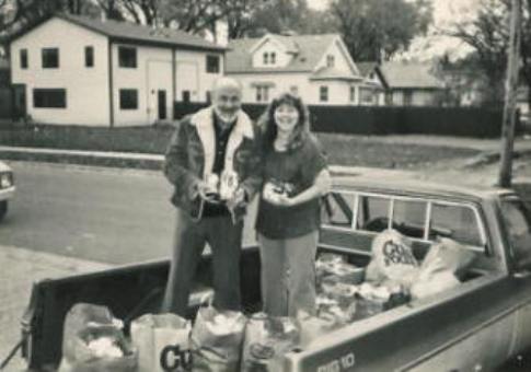 Pastor Paul and Judy, a member of the church, collect canned food for the 1st FoodDrive for the Grocery Shelf in 1983.